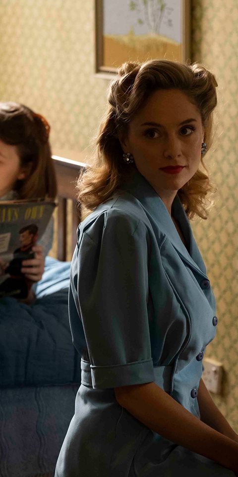Woman sitting on a chair with another woman reading a magazine on the bed behind her.