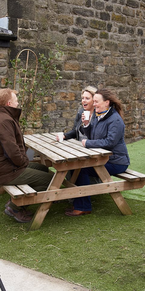 Three women sitting at a table outdoors with two cameramen surrounding them.